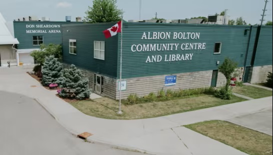 Albion Bolton Community Centre and Library building with a Canadian flag flying outside, featuring green siding and adjacent to Don Sheardown Memorial Arena.