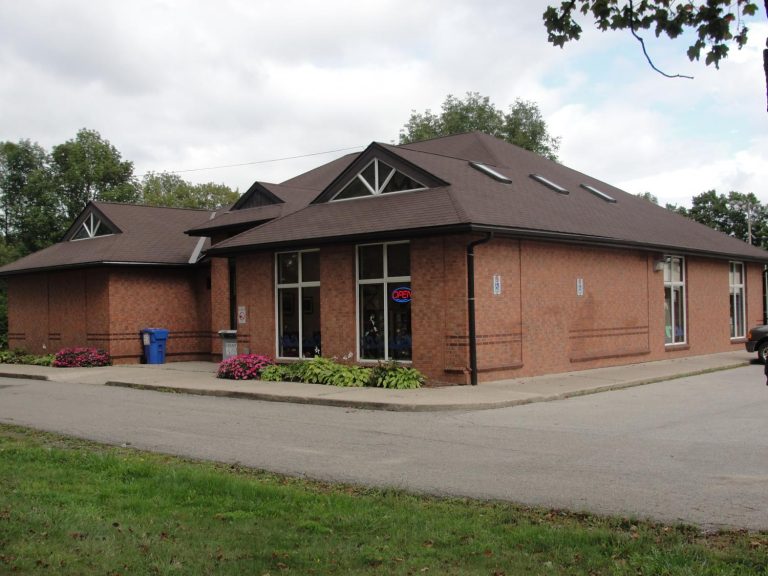 Exterior view of the Alton Library building. The structure is made of red brick with large windows, a sloping brown roof, and neatly maintained flower beds along the front entrance. A small sign reads 'Open,' and the surrounding area includes a paved driveway and green grass with some trees in the background.