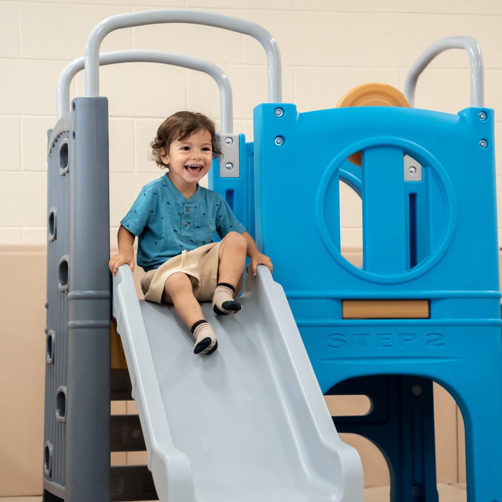 A little boy with a huge smile on a slide inside BrightStart on an inside plastic playset.