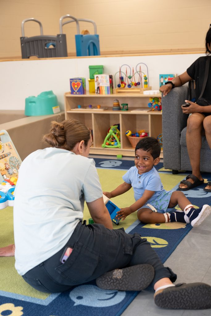A caregiver sits on the floor with a young child in a bright playroom, engaging in play with a toy house. The child, smiling brightly, interacts with the toy while another adult observes from a nearby couch. The playroom is filled with colourful toys and shelving, creating a welcoming environment at the BrightStart Caledon Family Centre for hands-on learning and joyful interactions.