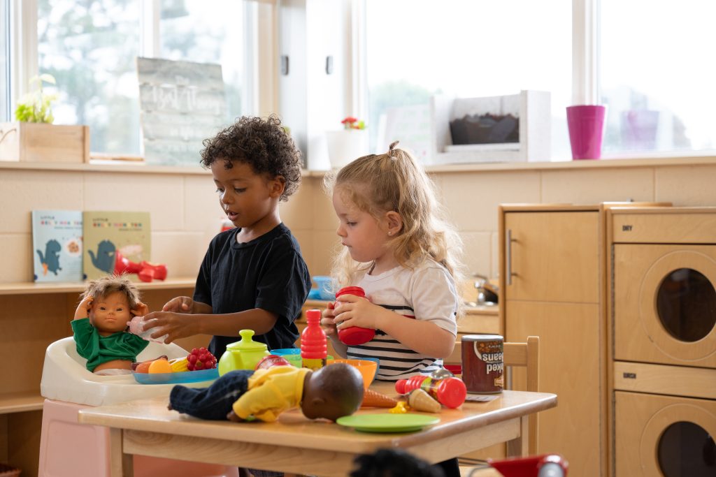 Two little preschoolers are playing house in a wooden kitchen with a baby doll in a highchair and all around a little play table with play food on it.