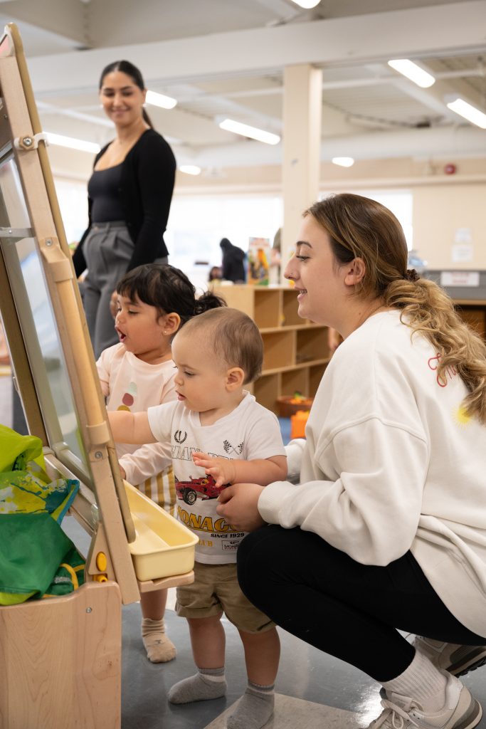 Two young women look at what two little toddlers are playing with on an easel in BrightStart.
