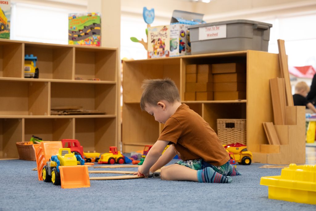 A young child sits on the floor in a playroom, focused on building with toy trucks and a wooden track. The child, dressed in a brown shirt and colourful shorts, is surrounded by various construction toys. Behind them, shelves are filled with toy trucks and building blocks, creating an inviting environment for creative play at the BrightStart Caledon Family Centre.