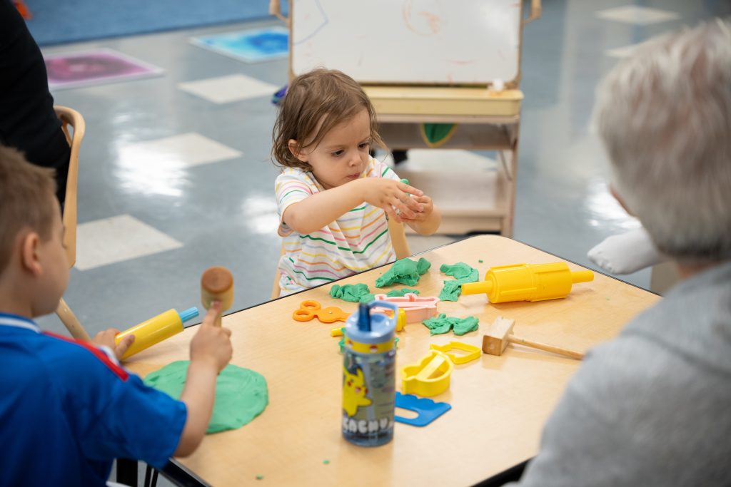 A young child, deeply focused, molds green playdough at a table, surrounded by various playdough tools, including a yellow rolling pin and orange scissors. Another child beside them is using a mallet to flatten the dough, while an adult watches from across the table. A Pikachu-themed water bottle is on the table, adding a playful element to the scene. The activity takes place in a vibrant, child-friendly space at the BrightStart Caledon Family Centre.