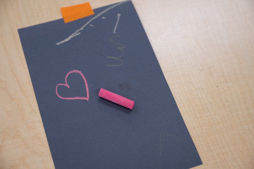 A black piece of construction paper is taped to a table at BrightStart with a pink piece of chalk and a drawing of a pink heart.