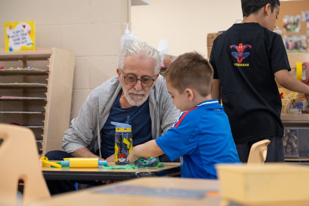 An older man with glasses and a grey hoodie sits at a table with a young child, guiding them in an activity involving playdough. The child, dressed in a blue shirt, focuses on their creative task, with a water bottle featuring Pikachu on the table. In the background, another child in a Spider-Man shirt is partially visible. The setting is a playroom at BrightStart Caledon Family Centre, where children are engaged in nurturing, hands-on activities.