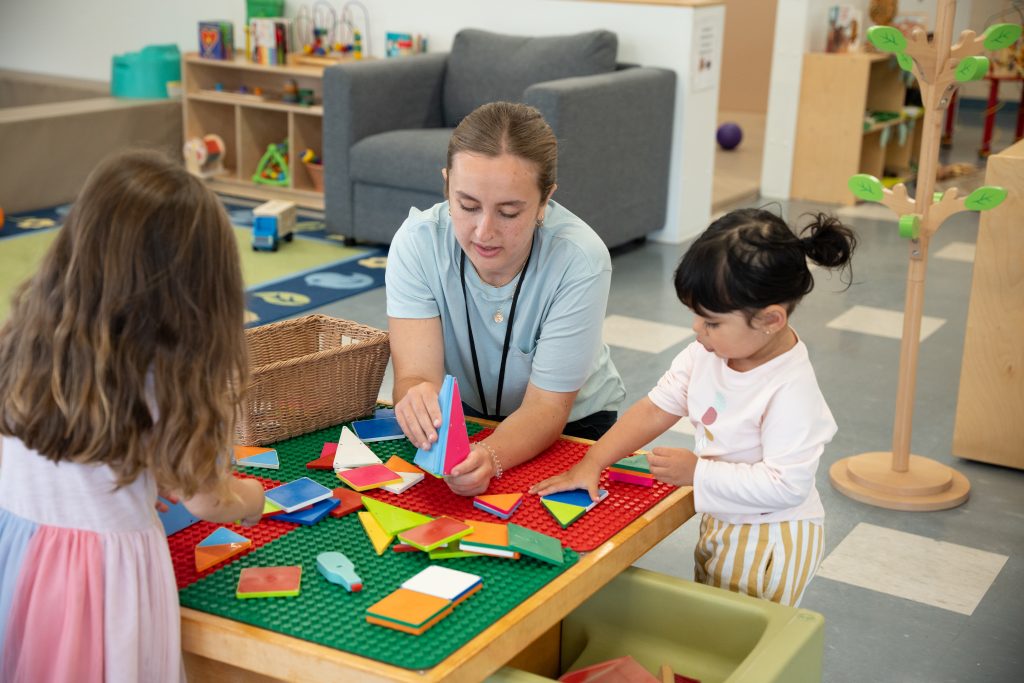 A caregiver kneels at a table with two young children, engaging them in an activity using colourful magnetic tiles. The children focus intently as they arrange the tiles on a large green board. The playroom at the BrightStart Caledon Family Centre is filled with toys and inviting spaces, providing a nurturing environment where children can explore hands-on learning.