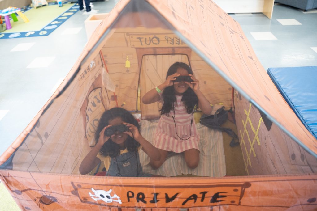 Inside a colorful play tent at BrightStart, two young girls are having fun. One girl, wearing overalls, is holding binoculars up to her eyes, while the other girl, dressed in a pink striped dress, is also using binoculars. The tent is decorated with playful designs, including a 'PRIVATE' sign, creating a sense of adventure and imagination.