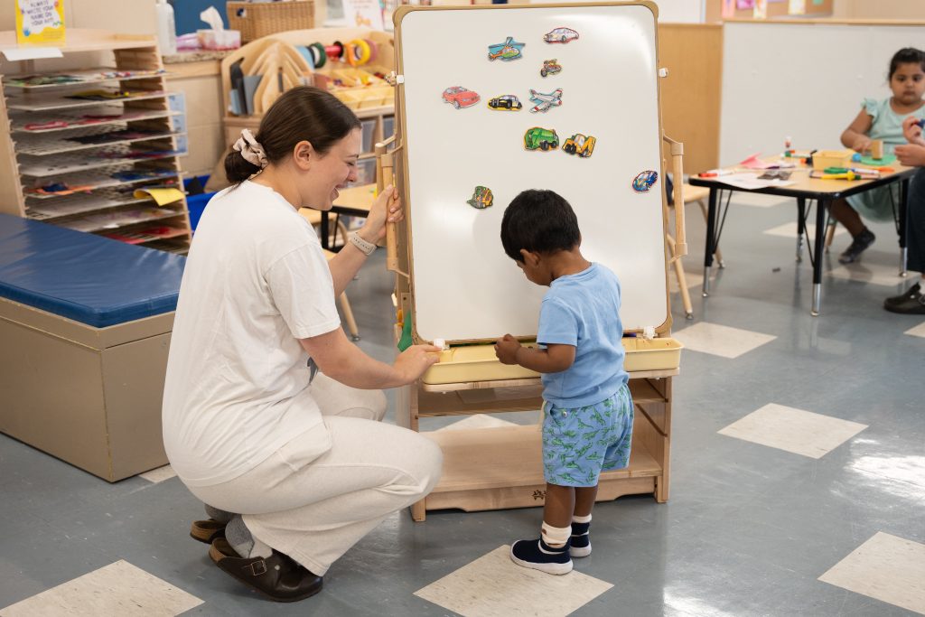 A caregiver is kneeling next to a young boy at BrightStart, assisting him with a magnetic board filled with various vehicle magnets. The boy, wearing a blue shirt and patterned shorts, is focused on placing a magnet while the caregiver, dressed in a white t-shirt and light pants, smiles encouragingly at him. In the background.