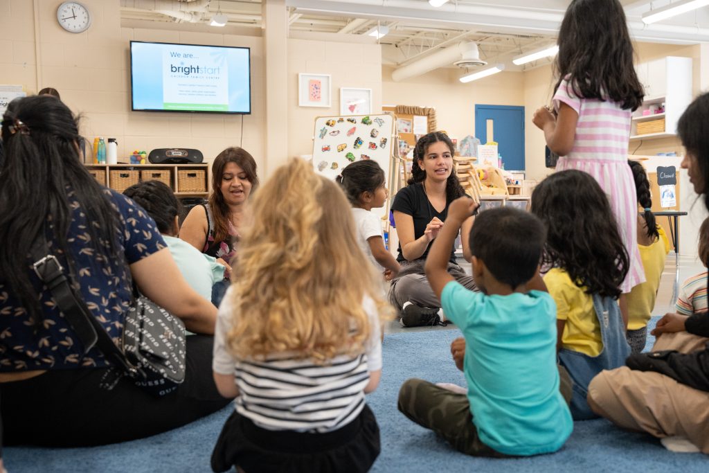 In a BrightStart family center, a group of children and adults are seated on the floor, engaged in a circle activity. Two staff members, one with long dark hair and the other with wavy brown hair, are animatedly interacting with the children. A television screen in the background displays the BrightStart logo. The atmosphere is lively and welcoming, highlighting the community-focused environment.