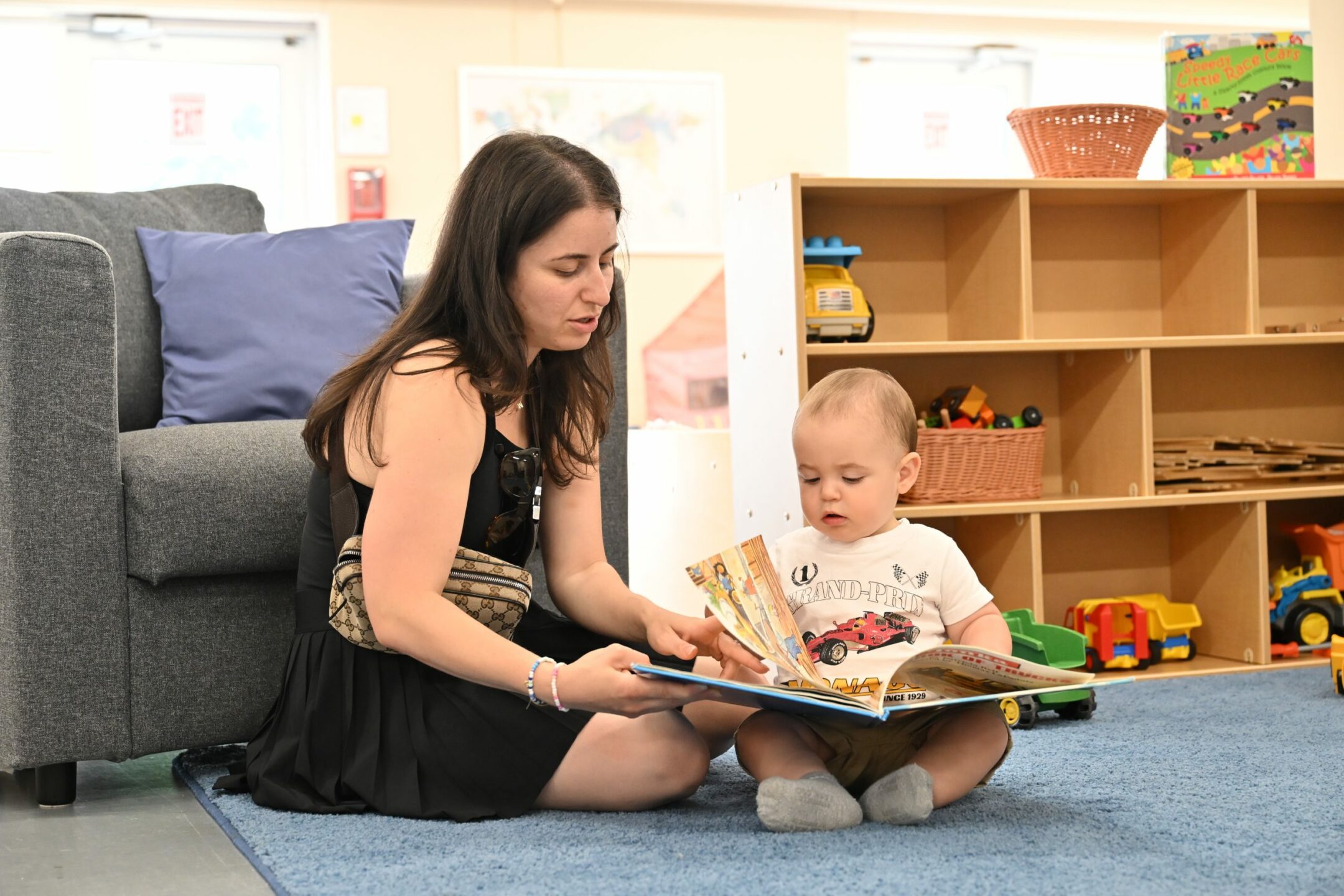 A woman sits on the floor with a young child, reading a picture book together in a cozy playroom. The child, dressed in a white shirt with a car print, focuses on the book while seated on a soft blue rug. In the background, shelves filled with toys and baskets add to the inviting atmosphere at the BrightStart Caledon Family Centre, encouraging literacy and bonding through reading.
