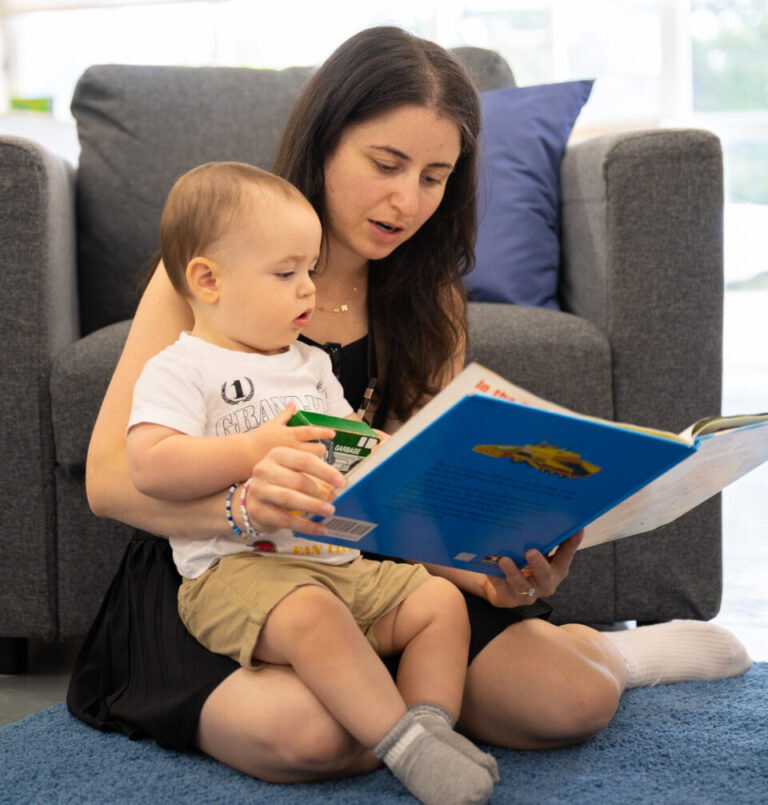 A baby sits on its mothers lap on a mat as a storybook is read