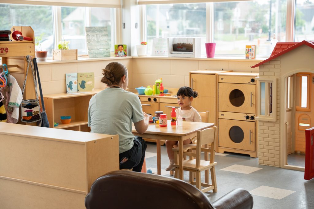 A caregiver and a young child sit at a small table in a bright playroom, engaging in a pretend tea party with toy food and kitchen items. The child smiles as they interact with the caregiver. Surrounding them are play kitchen appliances, a playhouse, and shelves filled with children's books and toys. The environment at the BrightStart Caledon Family Centre is designed to encourage imaginative play and nurturing interactions.