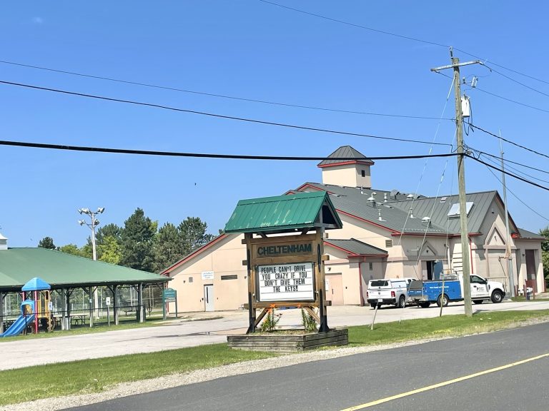 The Cheltenham Community Centre, a beige building with a green metal roof and red trim, is pictured with a large wooden sign out front that reads 'Cheltenham.' Below, the sign humorously states, 'People can't drive you crazy if you don't give them the keys.' In the foreground, there's a green-roofed pavilion next to a playground, and two white utility vehicles are parked in the community centre's lot. Power lines are visible across the image, and a bright blue sky stretches overhead.