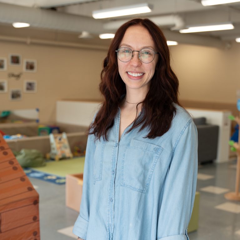Diane West, a staff member at BrightStart, is standing in a classroom environment, smiling and wearing glasses and a light denim shirt. The background includes various children's play items and furniture, creating a bright and welcoming learning space.