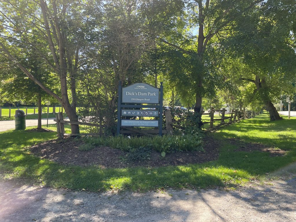 The entrance to Dick's Dam Park is marked by a blue-grey wooden sign that reads 'Dick's Dam Park - 250 Glasgow Road.' The sign is surrounded by trees and plants, and there is a gravel pathway leading into the park. A rustic wooden fence lines the edge of the park, adding to the natural, serene atmosphere. The park is lush with greenery, and the sunlight filters through the leaves, creating a peaceful, shaded area. In the background, more trees and open grassy areas are visible.