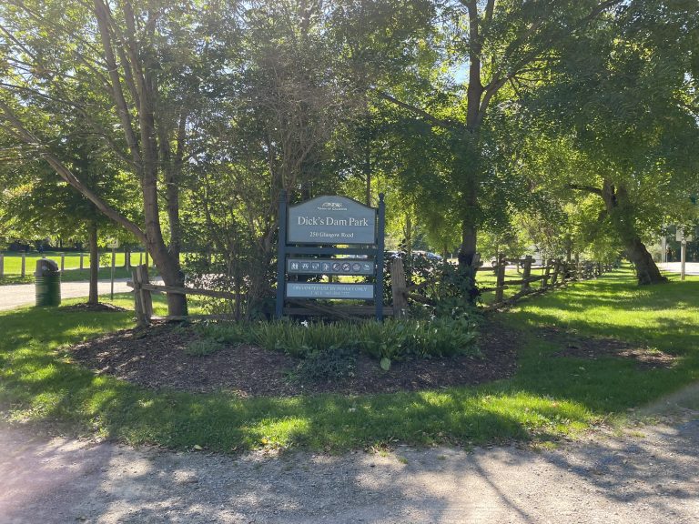 The entrance to Dick's Dam Park is marked by a blue-grey wooden sign that reads 'Dick's Dam Park - 250 Glasgow Road.' The sign is surrounded by trees and plants, and there is a gravel pathway leading into the park. A rustic wooden fence lines the edge of the park, adding to the natural, serene atmosphere. The park is lush with greenery, and the sunlight filters through the leaves, creating a peaceful, shaded area. In the background, more trees and open grassy areas are visible.