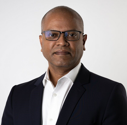 A professional headshot of Faizan Bari. He is wearing glasses and a dark suit with a white collared shirt, looking confidently at the camera. The background is a simple light grey.