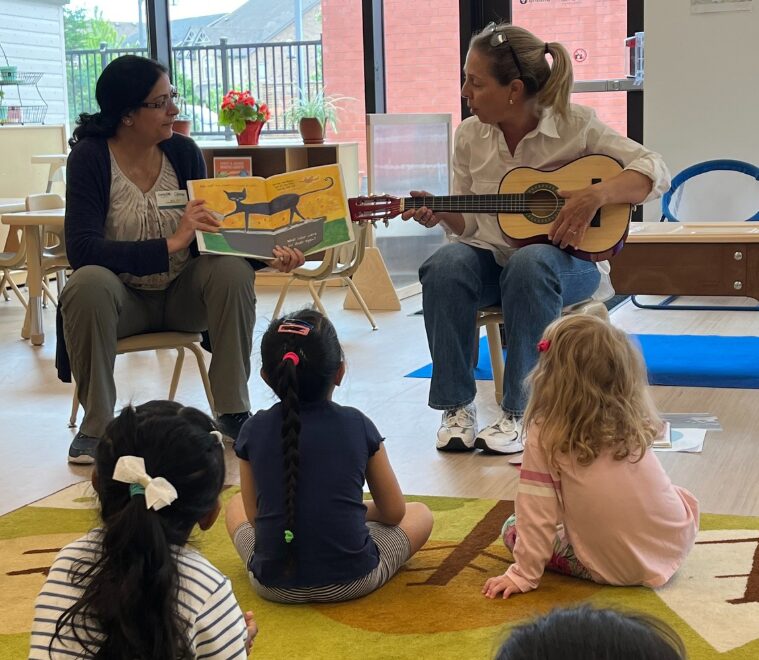 2 staff members sit in front of a group of preschool children; one plays a guitar and the other tells a story.