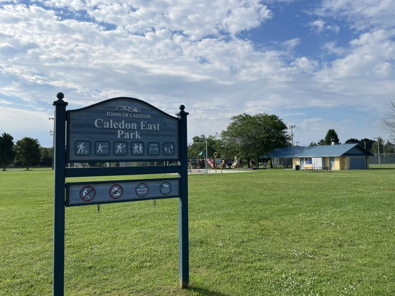 Caledon East Park sign in the foreground with grassy fields, a playground, and a pavilion visible in the background on a sunny day with scattered clouds.