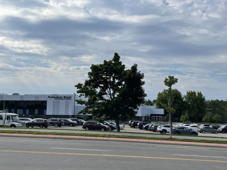 Exterior view of the Caledon East Community Complex on a partly cloudy day, with cars parked in the parking lot and trees in the foreground.