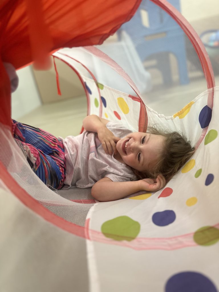 A young child is lying inside a colourful play tunnel, smiling and enjoying the moment. The tunnel is decorated with vibrant polka dots, creating a fun and playful atmosphere. The child appears relaxed and happy, engaging in active play at the BrightStart Caledon Family Centre.