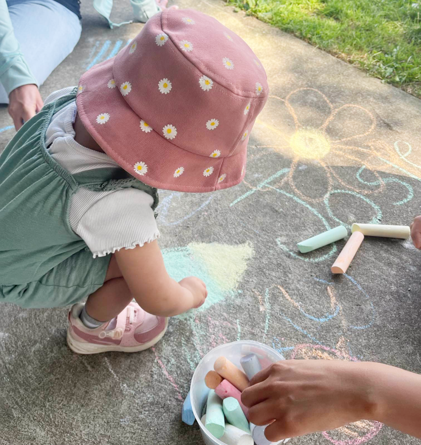A young child wearing a pink hat with daisy patterns is crouched on a sidewalk, drawing with colourful chalk. A bucket of chalk and various drawings, including a flower, are scattered on the pavement. The outdoor activity takes place in a park, encouraging creative expression through play at the BrightStart Caledon Family Centre.