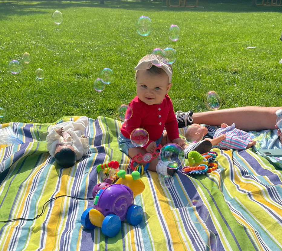 A baby wearing a red shirt and a pink headband sits on a colourful striped blanket outdoors, surrounded by toys and dolls. Bubbles float around the baby, adding a playful and joyful element to the scene. The grass and sunny environment create a relaxing and fun outdoor setting, capturing a moment of play at the BrightStart Caledon Family Centre.