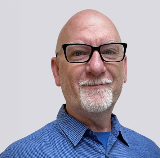 A professional headshot of John Heuer. He is wearing glasses and a blue checkered shirt, with a friendly smile. John has a bald head and a white beard. The background is light grey.