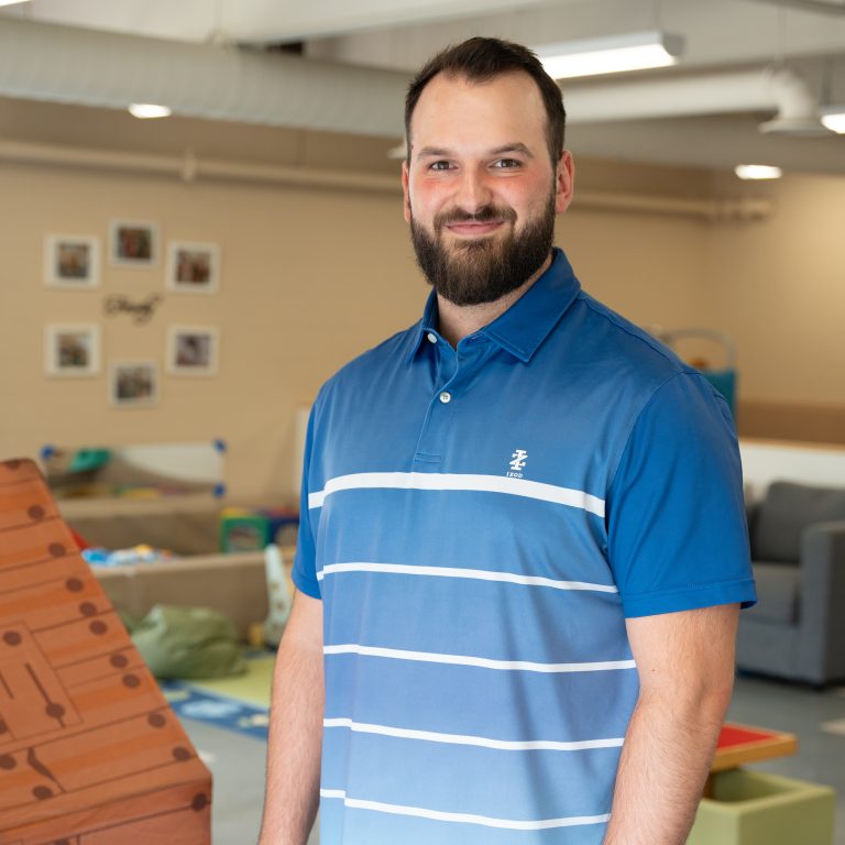 John-Paul Bertone, a staff member at BrightStart, is smiling and standing in a classroom setting. He is wearing a blue striped polo shirt with a relaxed and approachable demeanour. Behind him, the room is brightly lit with educational toys and furniture, creating a warm and engaging environment. The wall behind features a small gallery of family-themed framed pictures.