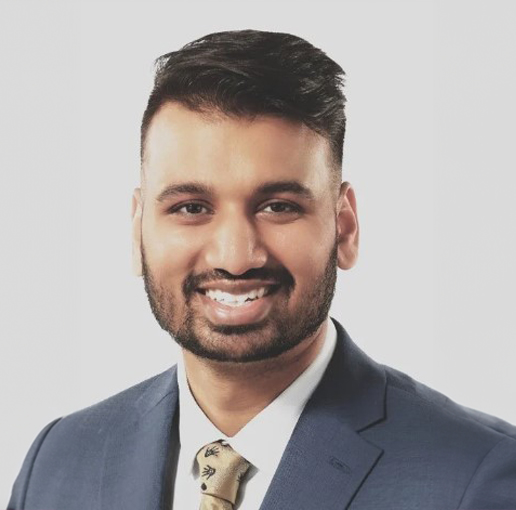 A professional headshot of Leo Rebello. He is smiling, wearing a dark blue suit, a white shirt, and a patterned beige tie. His hair is neatly styled, and he has a short beard. The background is a light grey, giving the image a polished and professional appearance.