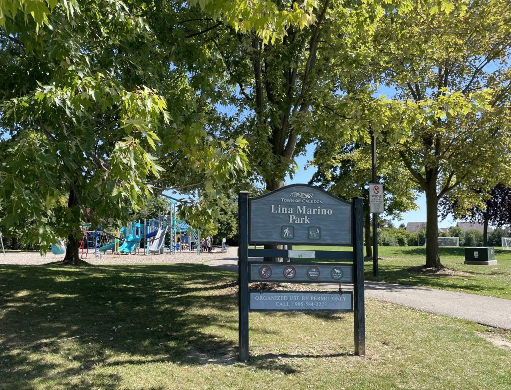 The entrance to Lina Marino Park is marked by a large blue-grey wooden sign that reads 'Lina Marino Park - Town of Caledon.' Behind the sign, a lush green park with a playground is visible, including blue slides and climbing structures. The park is shaded by large trees, casting a dappled shadow on the grass. A 'No Dogs Allowed' sign is placed near the entrance. In the background, a soccer field and more greenery stretch across the park, with a clear blue sky overhead.