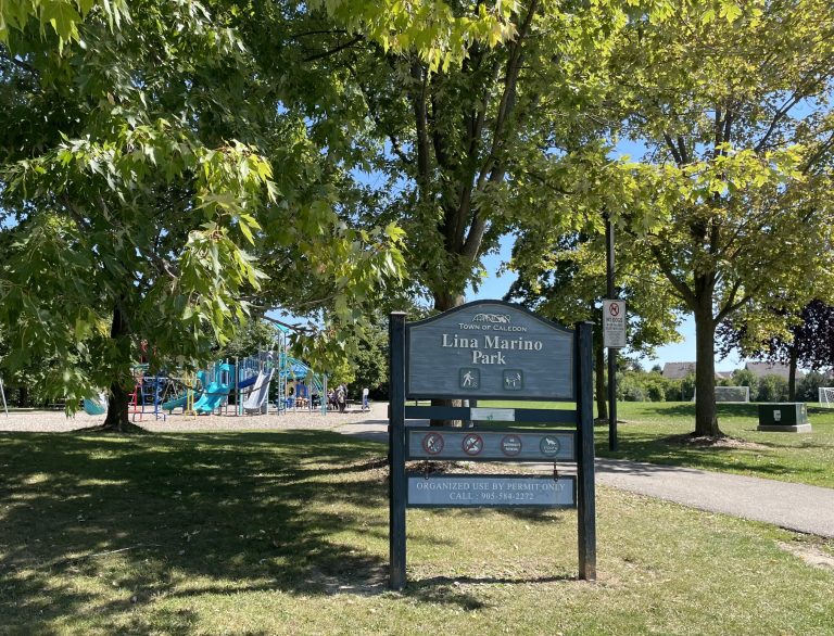 The entrance to Lina Marino Park is marked by a large blue-grey wooden sign that reads 'Lina Marino Park - Town of Caledon.' Behind the sign, a lush green park with a playground is visible, including blue slides and climbing structures. The park is shaded by large trees, casting a dappled shadow on the grass. A 'No Dogs Allowed' sign is placed near the entrance. In the background, a soccer field and more greenery stretch across the park, with a clear blue sky overhead.