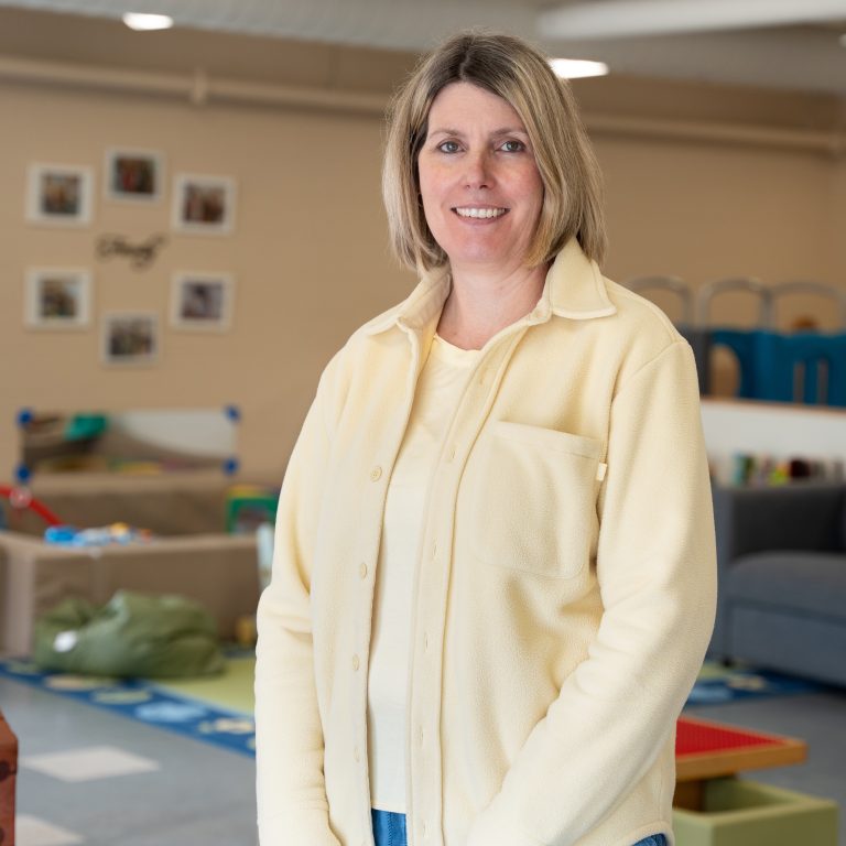 Lisa Chin, a staff member at BrightStart, smiles as she stands in a classroom setting. She is wearing a light yellow button-up shirt over a matching top. The background features children's toys and learning materials, with family-themed photos on the wall, creating a warm and engaging atmosphere.