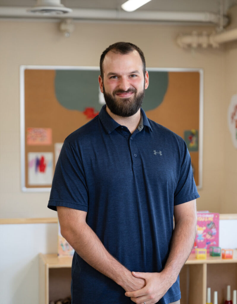 John-Paul Bertone, a staff member at BrightStart, is smiling and standing in a classroom setting. He is wearing a blue striped polo shirt with a relaxed and approachable demeanour. Behind him, the room is brightly lit with educational toys and furniture, creating a warm and engaging environment. The wall behind features a small gallery of family-themed framed pictures.
