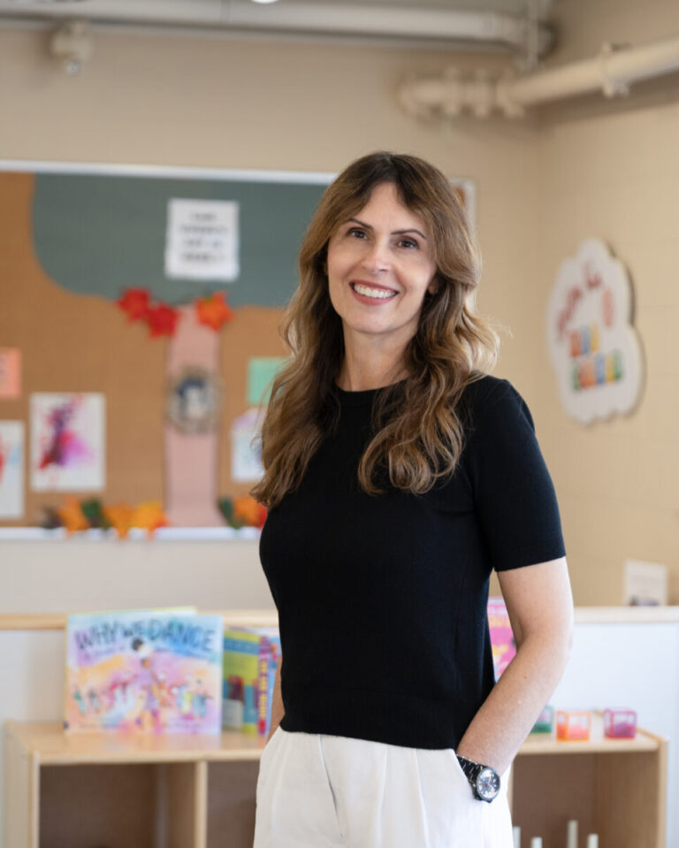 Teresa Colasanti, a staff member at BrightStart, is standing in a classroom setting, smiling and wearing a black short-sleeved shirt. The background features various children's play equipment and furniture, creating a cheerful and educational environment.