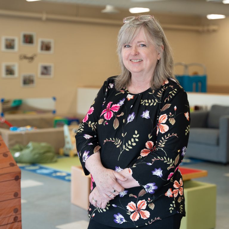 Shelly Ives, a staff member at BrightStart, is smiling and standing in a classroom setting. She is wearing a black top adorned with vibrant floral patterns in shades of pink, orange, and purple. The background features children's toys, learning materials, and framed family photos on the wall, giving a welcoming and educational environment.