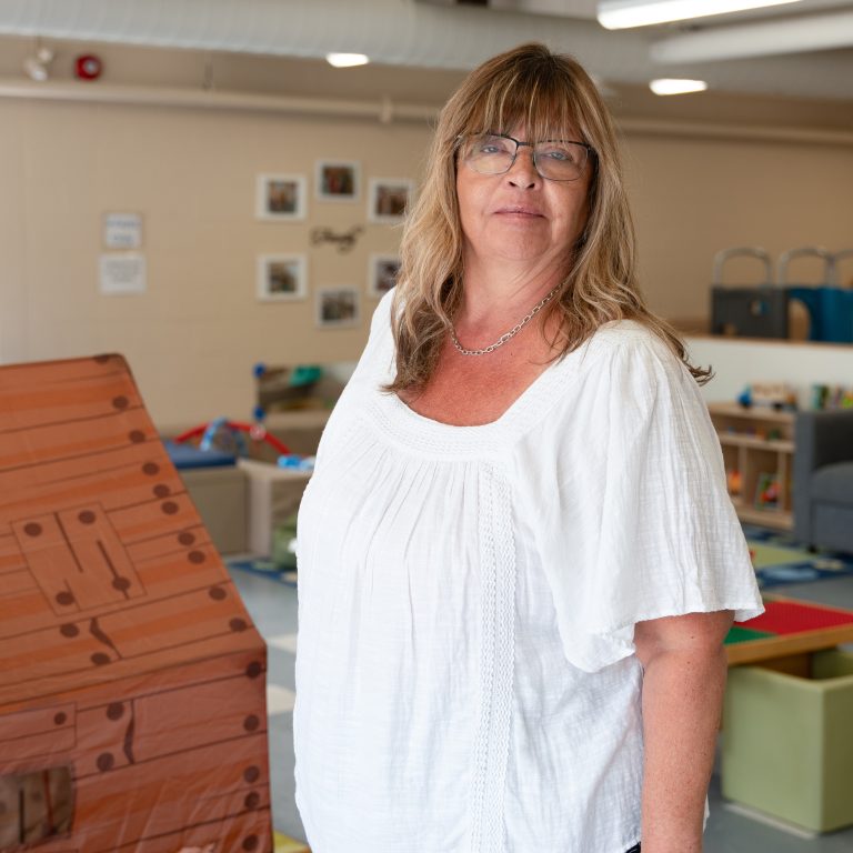 Susan Partyka, a staff member at BrightStart, stands in a classroom setting. She is wearing a white blouse with textured details and glasses, giving a confident yet approachable expression. The room behind her is set up with children’s toys and furniture, creating a warm and welcoming atmosphere, with family-themed photos on the wall.