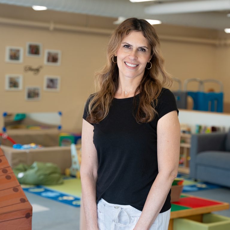 Teresa Colasanti, a staff member at BrightStart, is standing in a classroom setting, smiling and wearing a black short-sleeved shirt. The background features various children's play equipment and furniture, creating a cheerful and educational environment.