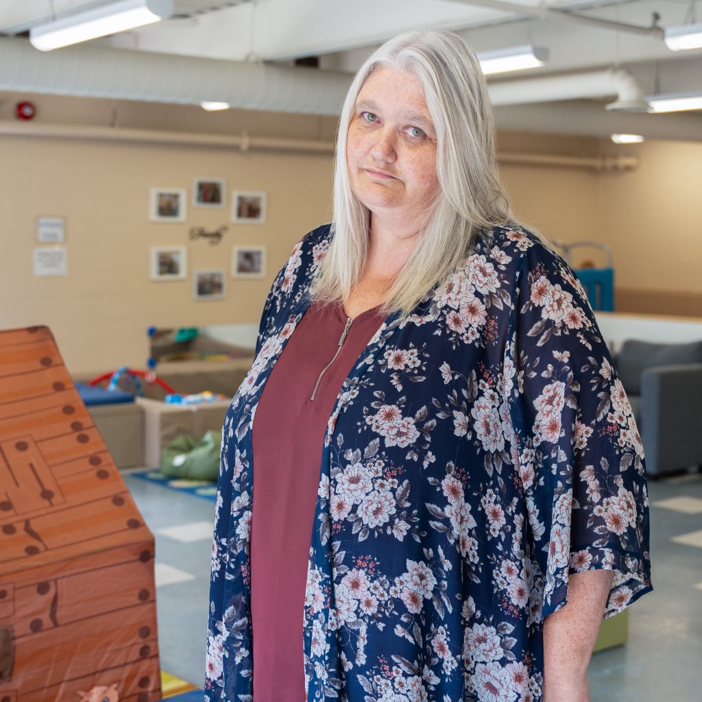Tracey Stewart, a staff member at BrightStart, stands in a classroom setting with a calm and friendly expression. She is wearing a maroon top and a navy floral cardigan. The room behind her is brightly lit, featuring children's educational toys and furniture, with family-themed framed pictures on the wall. The environment appears warm and welcoming.