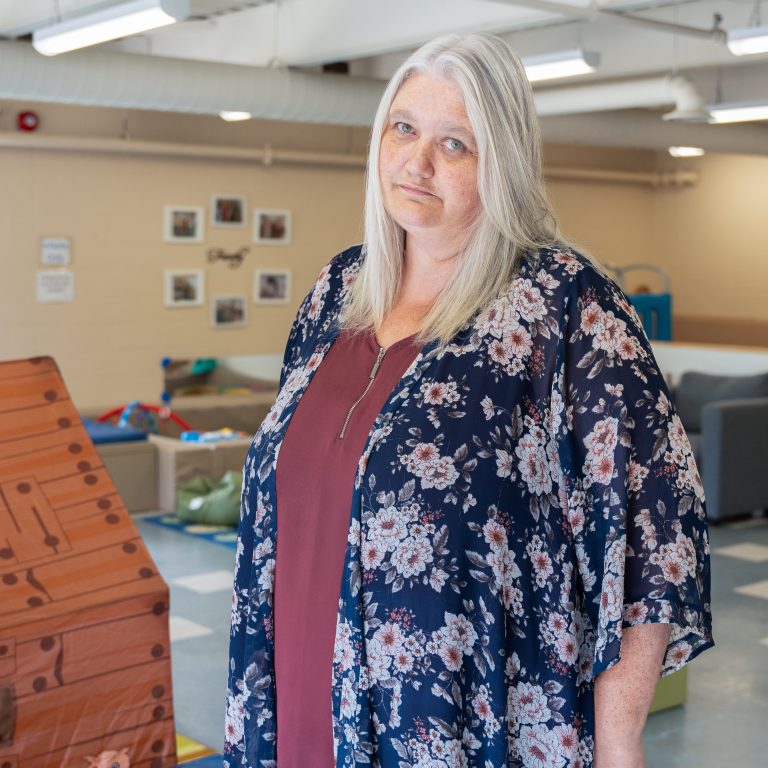 Tracey Stewart, a staff member at BrightStart, stands in a classroom setting with a calm and friendly expression. She is wearing a maroon top and a navy floral cardigan. The room behind her is brightly lit, featuring children's educational toys and furniture, with family-themed framed pictures on the wall. The environment appears warm and welcoming.