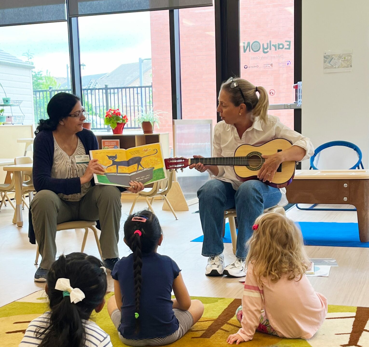 Two women lead a group of children in a music and storytime session. One woman holds a picture book open, showing an illustration of a cat, while the other woman plays an acoustic guitar. The children, seated on a colourful rug, listen attentively. The setting is a bright and welcoming room at the BrightStart Caledon Family Centre, encouraging literacy, music, and interaction in a nurturing environment.