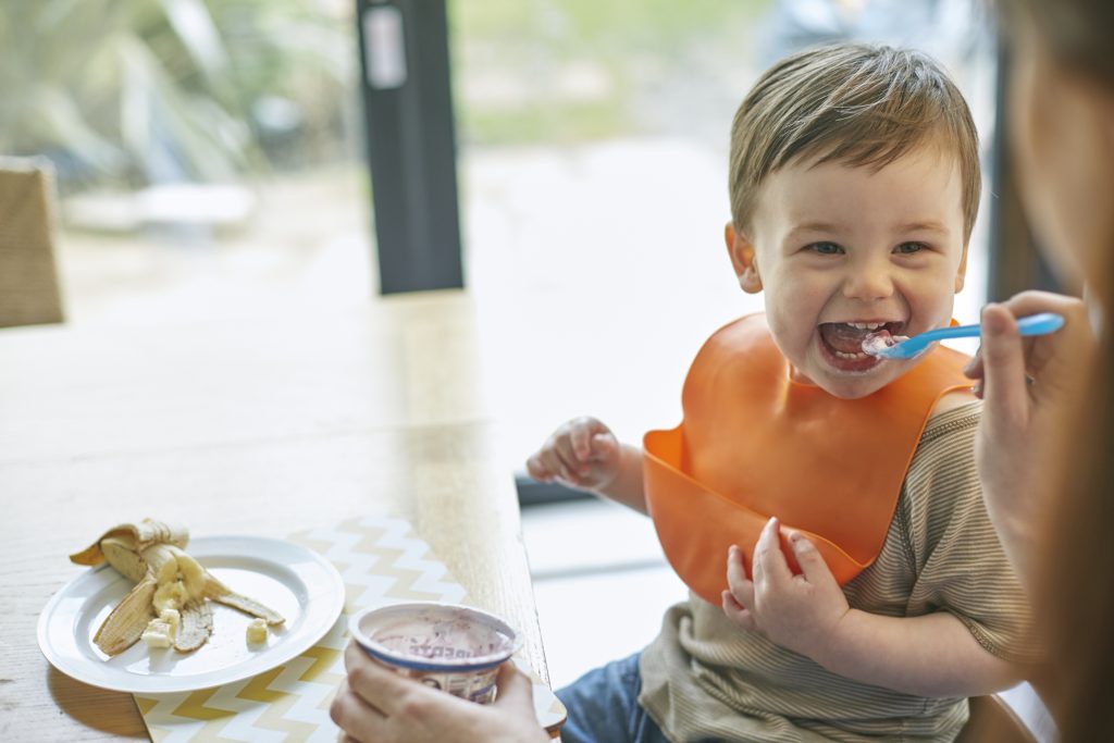 A happy baby boy sitting at a table being fed yogurt on a little blue spoon, while on a plate on the table was a banana.