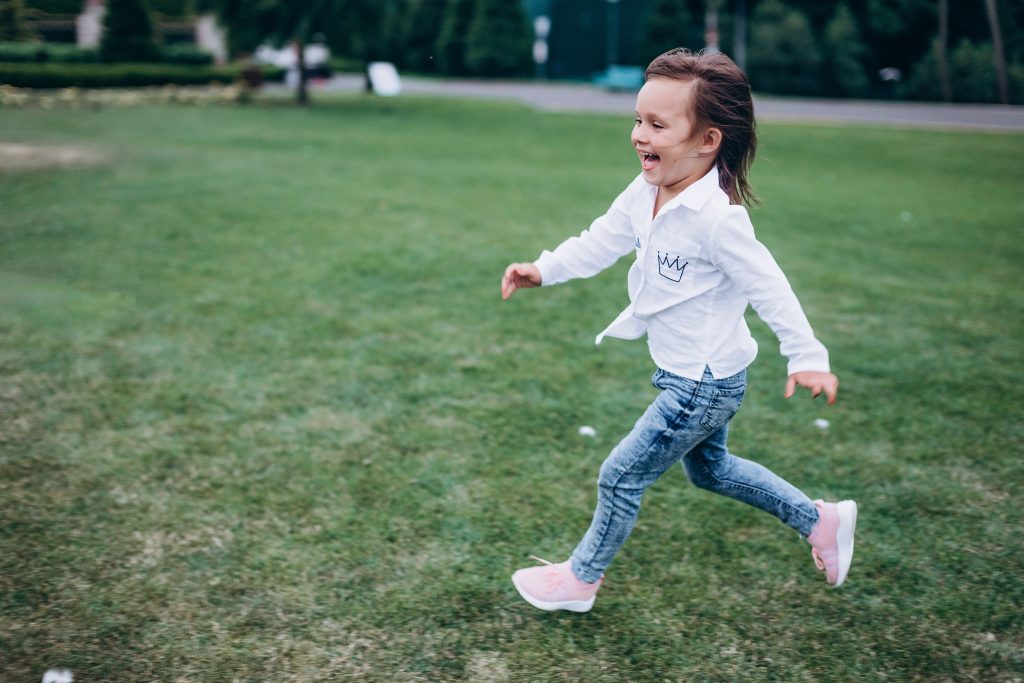 A happy child with brown hair, white shirt and jeans, is running and laughing on grass in a park.