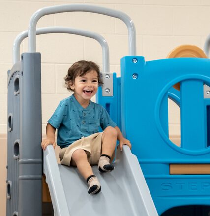 A young boy sits atop an indoor slide/playground, smiling at the camera