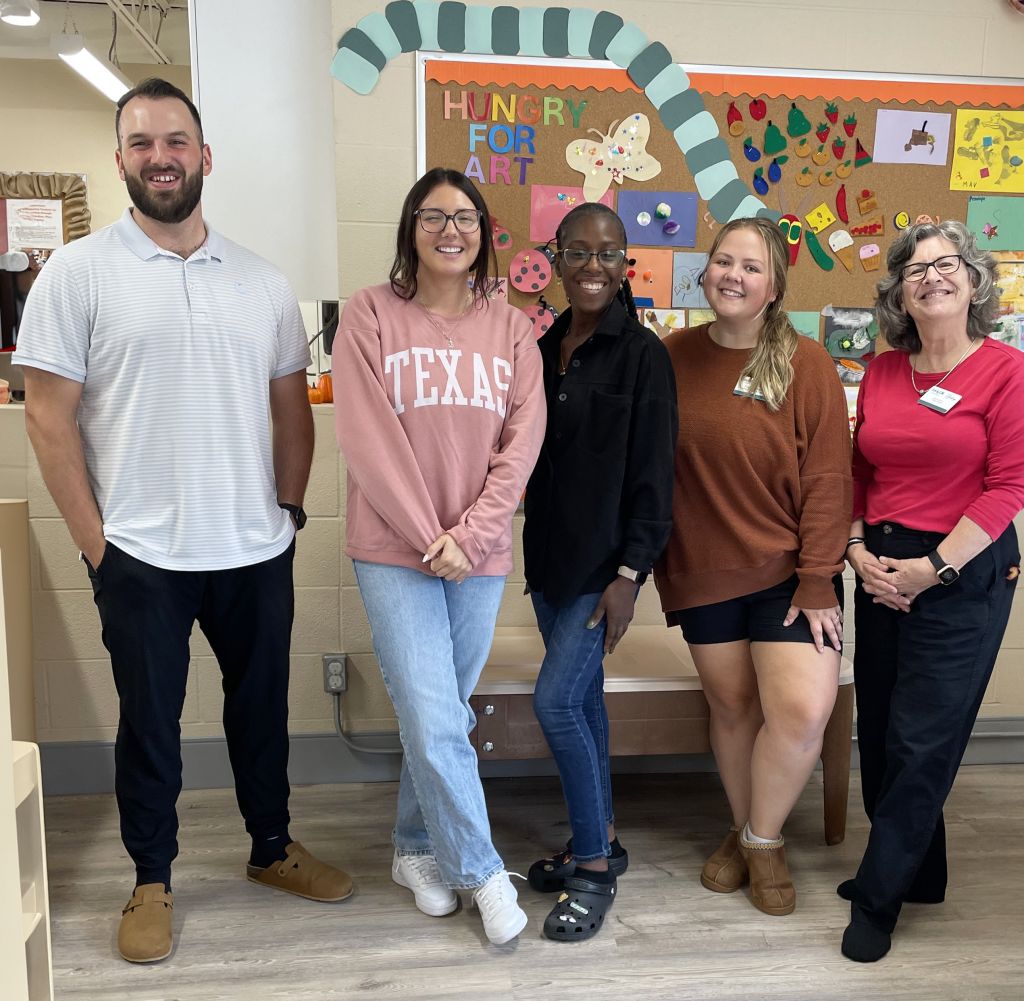 A group of five staff members stand together, smiling, in front of a colourful bulletin board titled 'Hungry for Art.' The group includes two men and three women, dressed casually, standing in a friendly and welcoming pose. The bulletin board behind them is decorated with various children's artwork and craft projects. The setting is bright and creative, reflecting the nurturing and artistic environment of the BrightStart Caledon Family Centre.