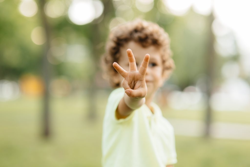 A young boy with curly hair playfully holds up three fingers while smiling at the camera. He is wearing a light yellow polo shirt and is set against a blurred background of greenery, indicating a sunny day in a park.